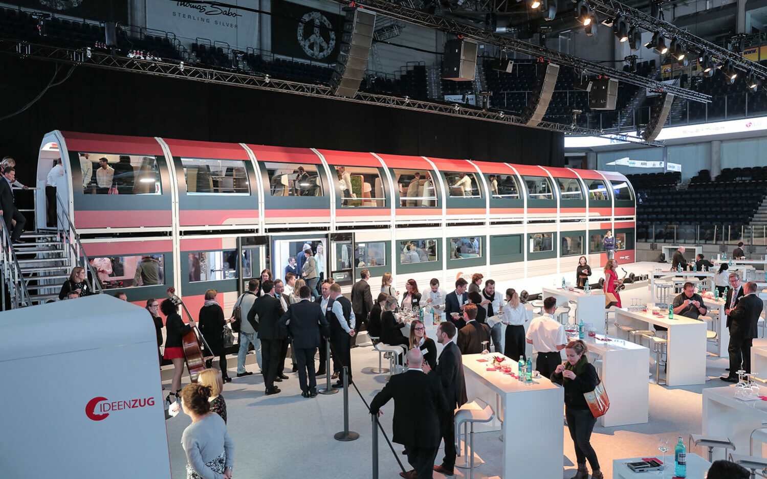 Auf dem Bild ist der DB Regio Ideenzug zu sehen. Vor dem Zug, als auch im Zug stehen Leute. Außerdem stehen vor dem Zug ein paar Stehtische, an denen sich Personen aufhalten.