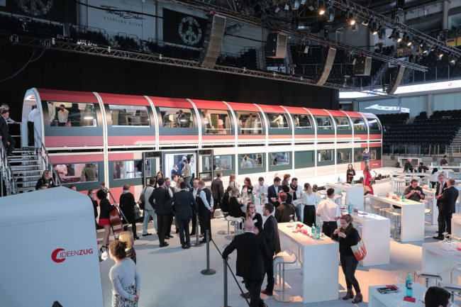 Auf dem Bild ist der DB Regio Ideenzug zu sehen. Vor dem Zug, als auch im Zug stehen Leute. Außerdem stehen vor dem Zug ein paar Stehtische, an denen sich Personen aufhalten.